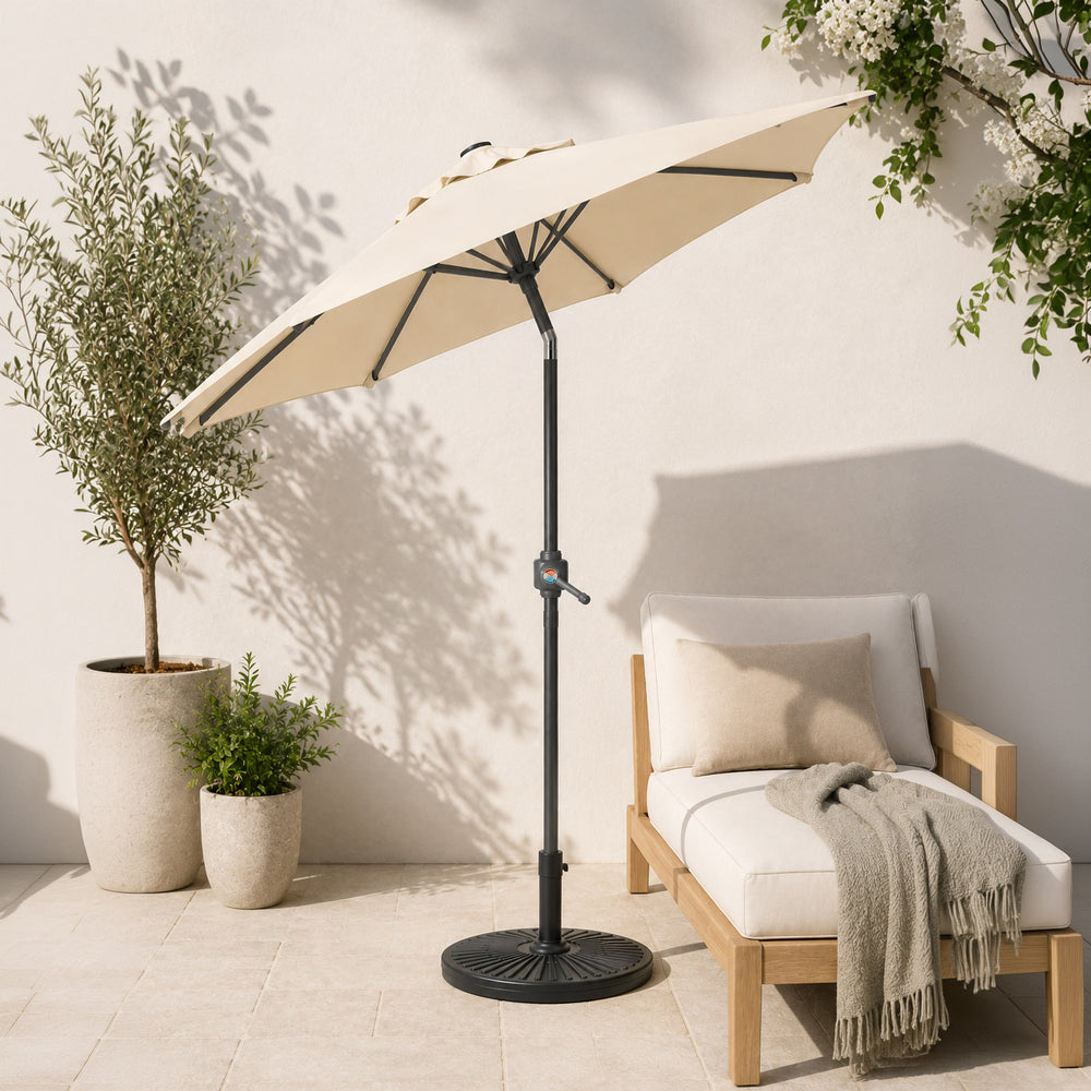 A beige patio umbrella with a black metal pole and base stands next to a white cushioned wooden lounge chair on a light stone patio. Two potted olive trees and climbing plants are visible against a white wall with soft shadows.