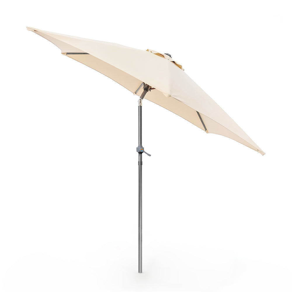 A beige patio umbrella with a black metal pole and base stands next to a white cushioned wooden lounge chair on a light stone patio. Two potted olive trees and climbing plants are visible against a white wall with soft shadows.