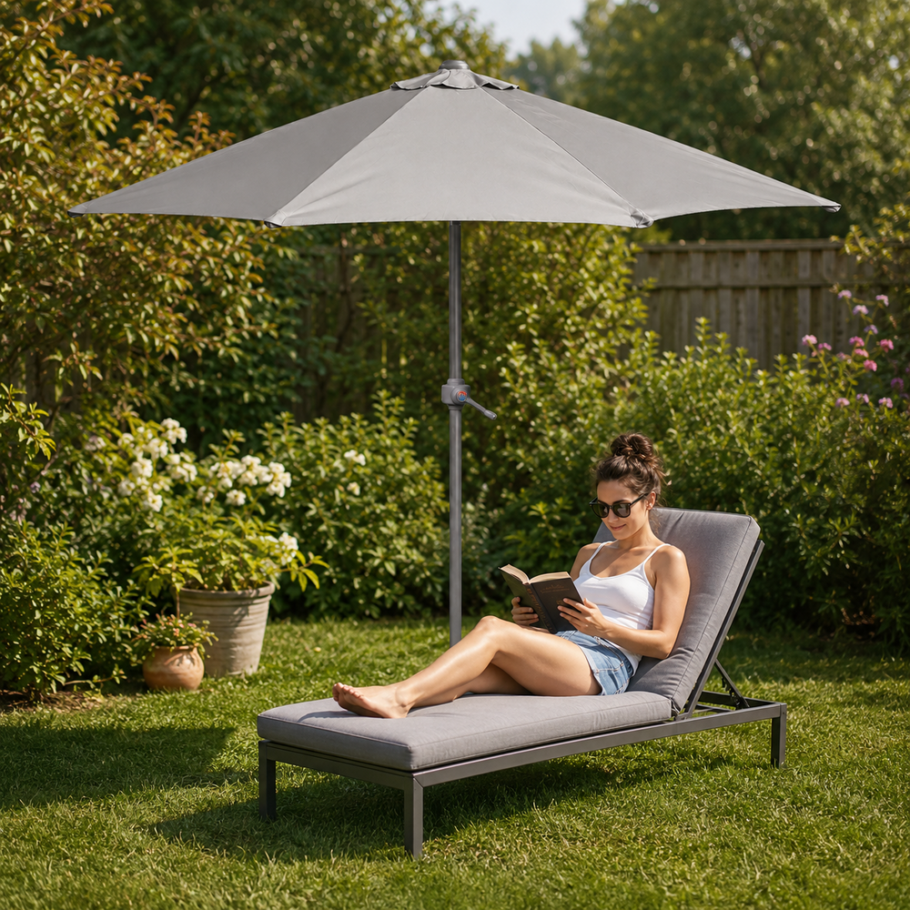 A woman relaxes on a gray outdoor chaise lounge under a matching patio umbrella in a lush green backyard garden.