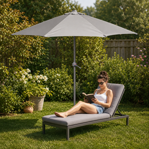 A woman relaxes on a gray outdoor chaise lounge under a matching patio umbrella in a lush green backyard garden.