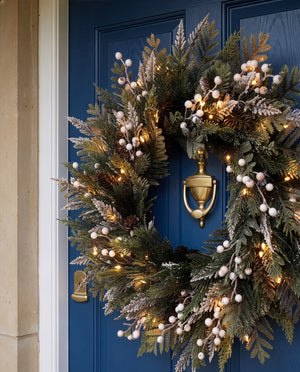 A festive holiday wreath with green pine branches, white berry clusters, and warm white string lights, hung on a deep blue front door with a brass knocker.