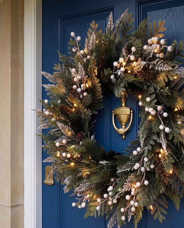 A festive holiday wreath with green pine branches, white berry clusters, and warm white string lights, hung on a deep blue front door with a brass knocker.