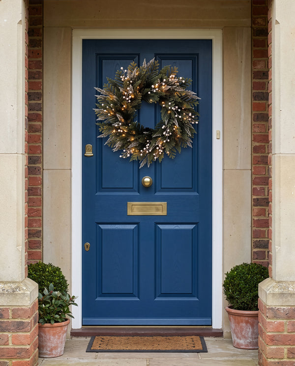 A classic navy blue front door with brass hardware, decorated with a green pine wreath featuring white berries and warm string lights, set in a brick and stone entryway with potted plants.