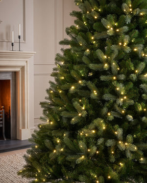 A close-up of a green Christmas tree decorated with warm white string lights, set in a cozy living room next to a fireplace.