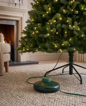 A green electric Christmas tree humidifier and light controller sits on a beige textured rug beneath a lit Christmas tree in a cozy living room with a fireplace in the background.