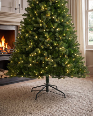 A green artificial Christmas tree with warm white string lights, standing on a black metal stand in a cozy living room setting with a fireplace and window in the background.