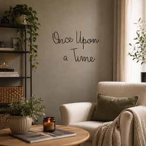 A cozy living room scene with a cream armchair, green pillow, knit throw blanket, potted plants, a black shelving unit, and a round wooden coffee table. On the wall, black script text reads 'Once Upon a Time'.
