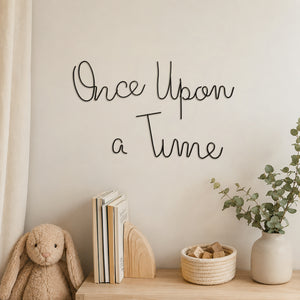 Black metal script wall art reading 'Once Upon a Time' mounted on a beige wall above a wooden shelf styled with a plush bunny, books, a small basket, and a potted eucalyptus plant.