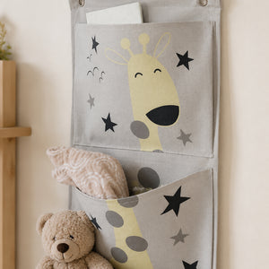 A light gray fabric hanging organizer with three pockets, printed with a smiling giraffe and black stars. The top pocket holds a white card, the middle pocket contains a soft pink blanket, and the bottom pocket is partly covered by a beige teddy bear.