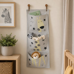 A gray fabric hanging organizer with three compartments, decorated with cartoon giraffes, stars, a monkey, and a lion, shown hanging on a beige wall in a styled nursery setting.