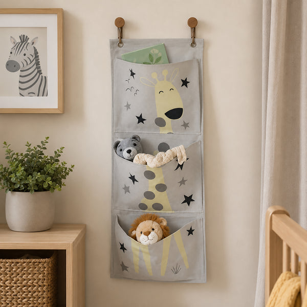 A gray fabric hanging organizer with three compartments, decorated with cartoon giraffes, stars, a monkey, and a lion, shown hanging on a beige wall in a styled nursery setting.
