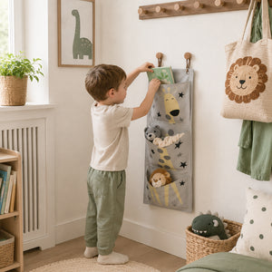 A young child stands in a nursery, reaching into a gray fabric hanging organizer with animal pockets and star details. The room has a white wall, a wooden coat rack, a potted plant, and a woven tote bag with a lion face.