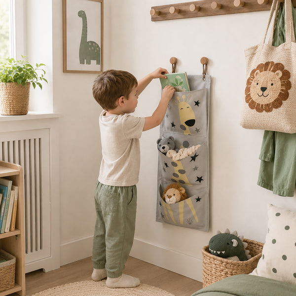 A young child stands in a nursery, reaching into a gray fabric hanging organizer with animal pockets and star details. The room has a white wall, a wooden coat rack, a potted plant, and a woven tote bag with a lion face.