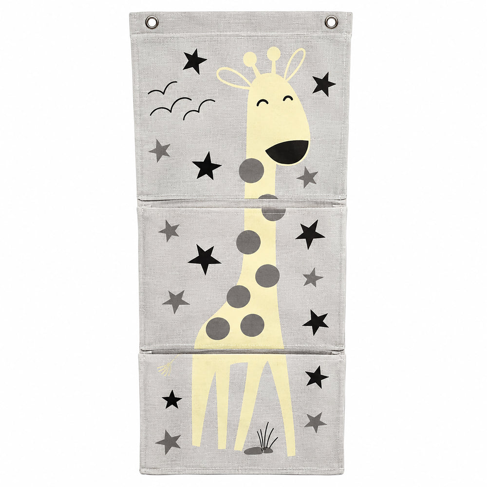 A gray fabric hanging organizer with three front pockets printed with a yellow giraffe and black stars, hanging on a beige wall in a styled room with a wooden shelf, framed art, and a small plant.