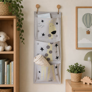 A gray fabric hanging organizer with three front pockets printed with a yellow giraffe and black stars, hanging on a beige wall in a styled room with a wooden shelf, framed art, and a small plant.