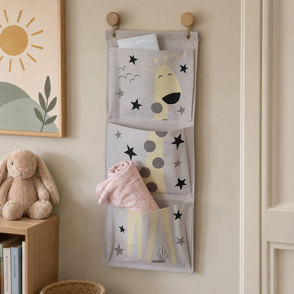 A three-pocket fabric hanging organizer with a giraffe and star print, hung on a beige wall in a nursery-like room. A plush bunny sits on a wooden shelf beside it, and folded blankets are tucked into the pockets.