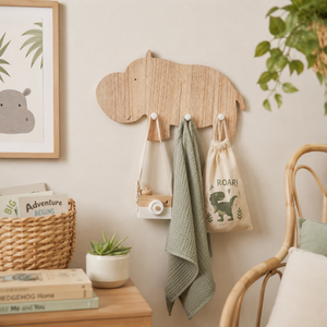 A light natural wood hippo-shaped wall hook rack mounted on a beige wall, holding a cream tote bag, a sage green textured towel, and a small wooden camera. The scene is styled as a nursery or children's room with a framed hippo print, a wicker basket, books, a small potted succulent, and a rattan chair.