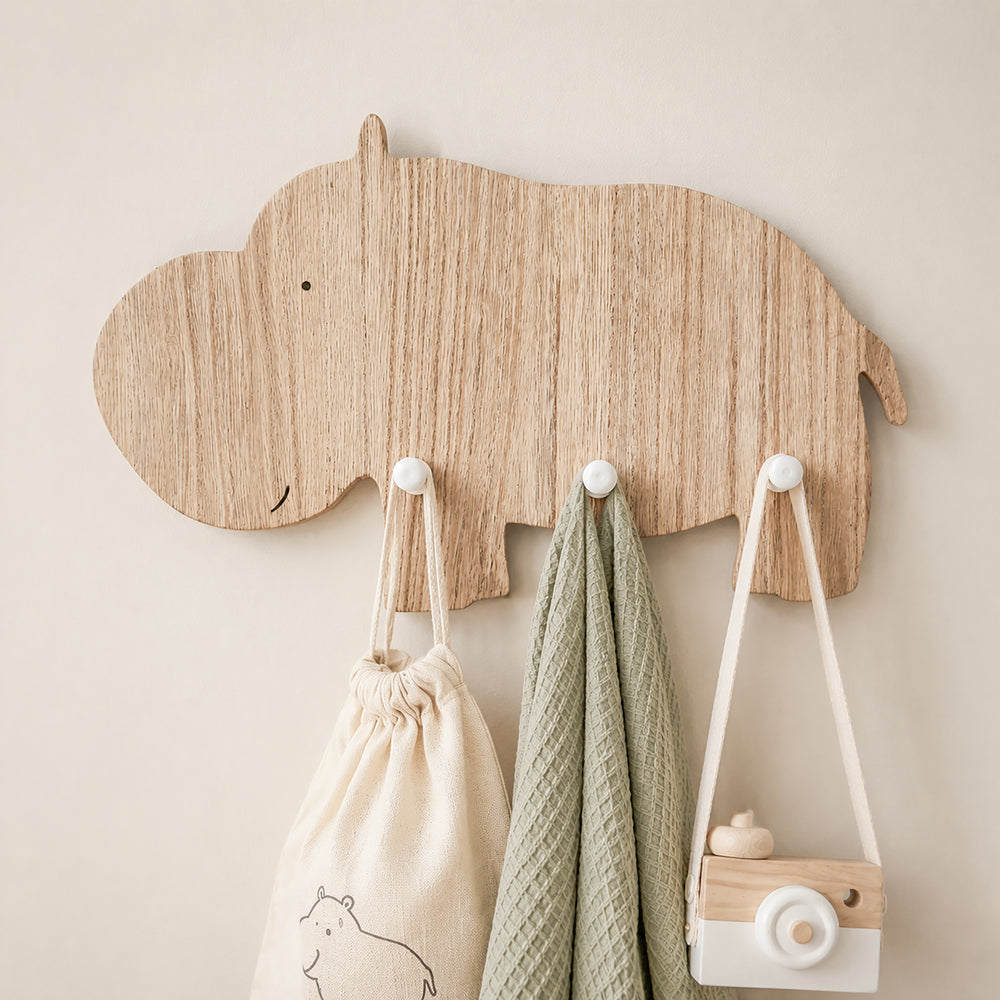 A light natural wood hippo-shaped wall hook rack mounted on a beige wall, with three hanging accessories: a cream drawstring bag, a sage green textured cloth, and a small wooden camera bag.