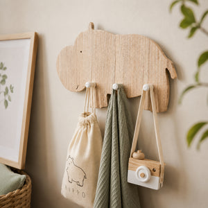 A light natural wood hippo-shaped wall hook rack mounted on a beige wall, holding a cream drawstring bag, a green textured towel, and a small wooden camera accessory.