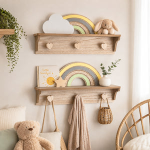 Two rustic wooden floating shelves mounted on a beige wall in a softly styled nursery. The shelves hold decorative items including a white cloud plaque, a wooden rainbow, a plush bunny, a small book, a star ornament, a vase with greenery, and hanging rope hooks with a beige towel and woven basket.