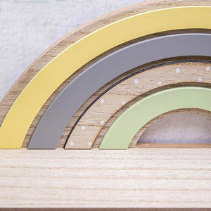 A close-up of a wooden rainbow toy with layered arches in yellow, gray, beige, and pale green, some with small white dot details, photographed on a light wood surface.