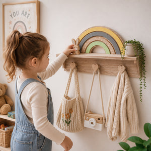 A young child in denim overalls reaches toward a wooden wall-mounted shelf with heart-shaped hooks, a rainbow decoration, a knitted tote bag, a small camera, and a cream knit sweater in a bright nursery setting.