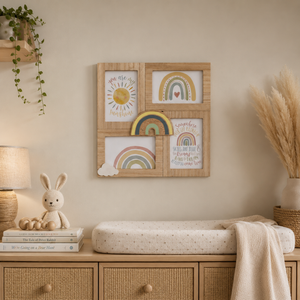 A styled nursery wall with a four-panel wooden frame holding pastel rainbow and sun illustrations, mounted above a changing table with a cream cushion and blanket. A lamp, books, a plush bunny, dried grass, and a hanging plant are visible in the room.