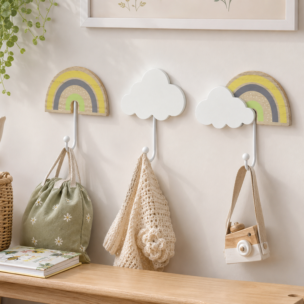 Three wall hooks styled in a nursery or children's room setting, featuring two wooden rainbow designs and one white cloud design. The hooks are mounted on a light beige wall above a wooden shelf, holding a sage green drawstring bag, a beige crochet bag, and a small wooden toy camera.