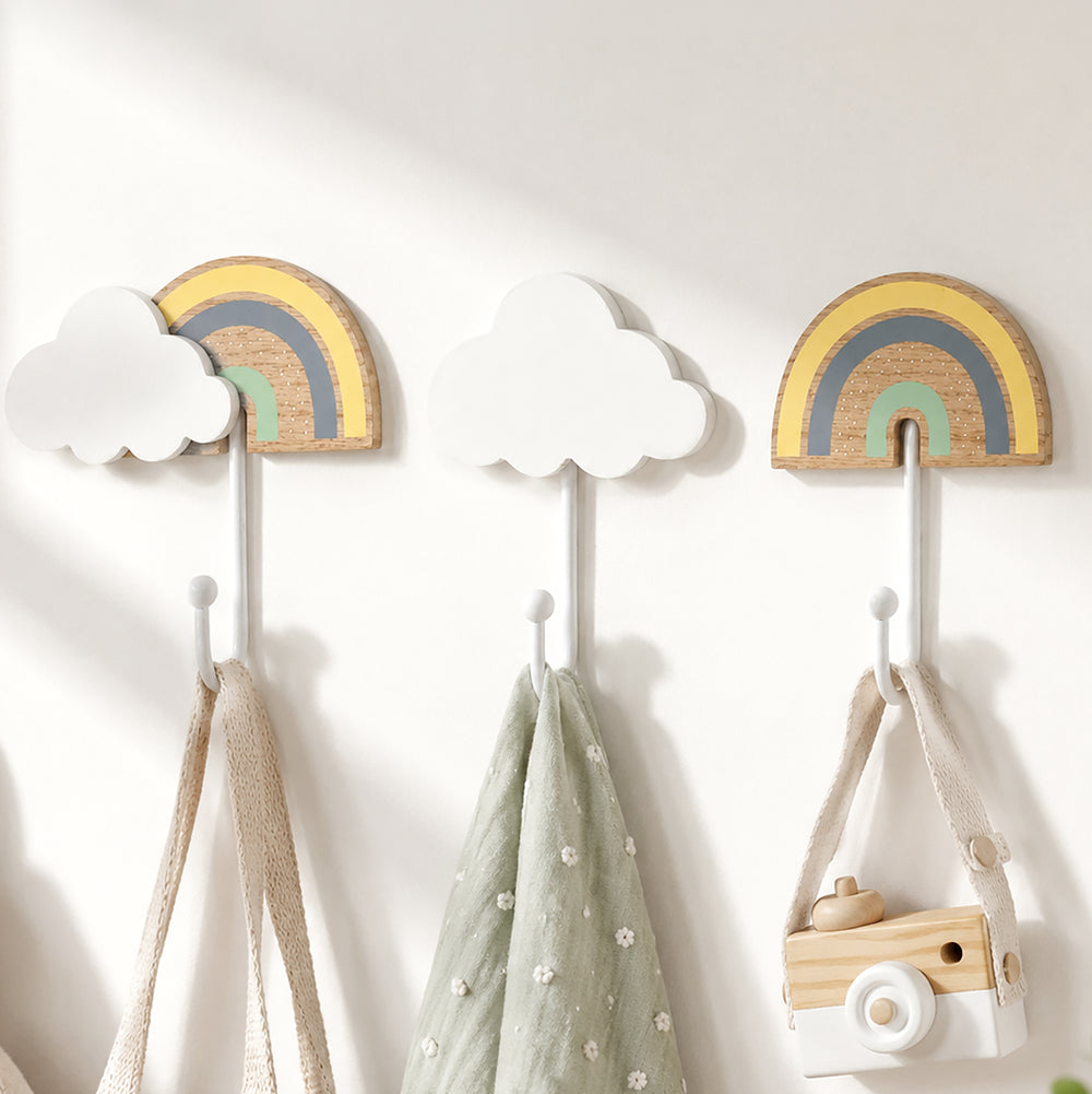 Three wall-mounted children's coat hooks with cloud and rainbow designs in soft pastel colors, shown hanging bags and a toy camera against a white wall.