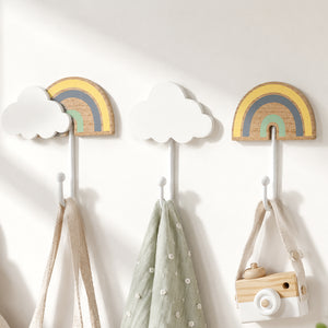 Three wall-mounted children's coat hooks with cloud and rainbow designs in soft pastel colors, shown hanging bags and a toy camera against a white wall.