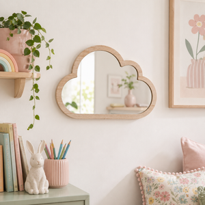 A cloud-shaped wooden wall mirror with a natural light wood frame, hanging on a white wall in a styled nursery or children's room. The scene includes a green trailing plant on a shelf, pastel books, a white bunny figurine, a pink pencil cup, and a floral pillow.