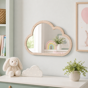 A cloud-shaped wooden wall mirror with a natural light wood frame hangs on a pale blue-gray wall above a white dresser. The scene includes a plush bunny, a small cloud-shaped lamp, a potted plant, shelving, and framed wall art, suggesting a styled nursery or children's room setting.