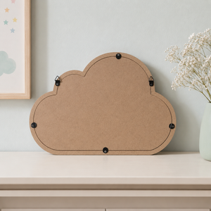 A cloud-shaped cork board with black metal clips on the back, displayed on a light beige shelf against a pale blue-gray wall, with a vase of baby's breath and a framed print partially visible.