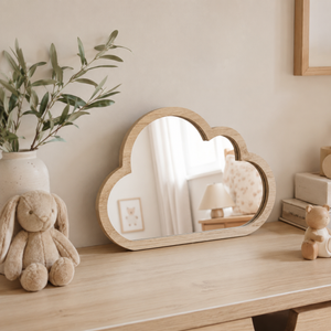 A cloud-shaped wooden mirror with a natural light oak frame sits on a light wood table against a beige wall. The scene is styled with a plush bunny toy, a white vase with green leaves, and a small wooden teddy bear.