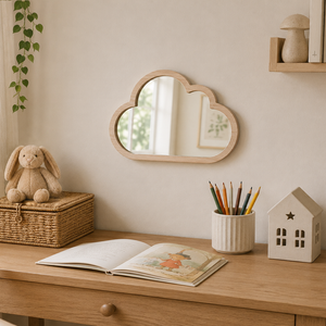 A cloud-shaped wooden wall mirror hangs on a beige wall above a light wood desk styled with a bunny plush, woven basket, open book, pencil cup, and a small house-shaped decor piece.