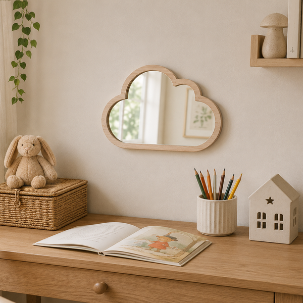A cloud-shaped wooden wall mirror hangs on a beige wall above a light wood desk styled with a bunny plush, woven basket, open book, pencil cup, and a small house-shaped decor piece.