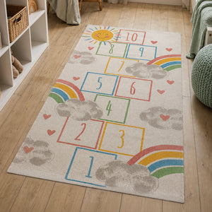 A rectangular children's area rug with a colorful hopscotch-style number grid, rainbows, clouds, hearts, and a smiling sun, shown on a light wood floor in a nursery-like room.