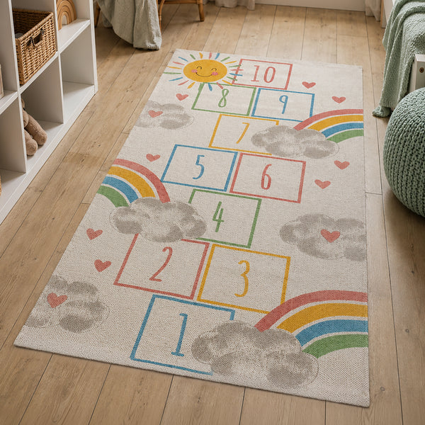 A rectangular children's area rug with a colorful hopscotch-style number grid, rainbows, clouds, hearts, and a smiling sun, shown on a light wood floor in a nursery-like room.