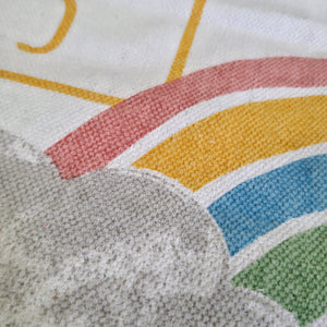 Close-up of a textured fabric print featuring a partial rainbow with pink, yellow, blue, and green arcs over a white background with a gray cloud.