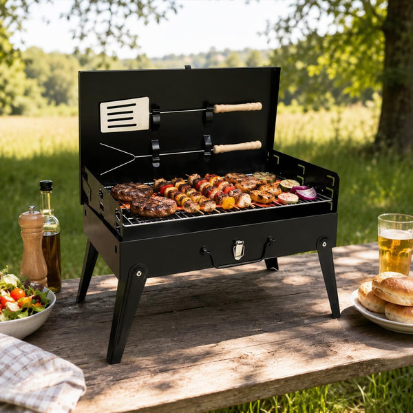 A black portable folding grill sits on a wooden picnic table outdoors, with meat, kebabs, and vegetables cooking on the grate. A spatula and fork hang from the open lid, and a salad, bottles, and bread are placed on the table beside it.