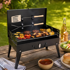 A black foldable mini charcoal grill with a lid, shown open on a wooden outdoor table with food cooking inside. The grill has two utensils hanging from the lid with light wood handles.