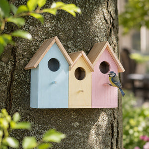 Three small house-shaped birdhouses in pastel blue, cream, and pink are mounted on a tree trunk outdoors, with a small bluebird perched on the right birdhouse.