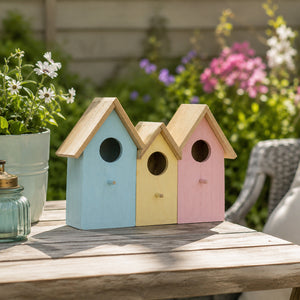 Three small wooden birdhouses in pastel blue, yellow, and pink, arranged on a weathered wooden table in a garden setting with blurred flowers and patio furniture in the background.