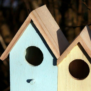 Two small wooden birdhouses with pitched roofs and round entry holes, shown outdoors against a blurred dark background. One is painted light blue and the other pale yellow.