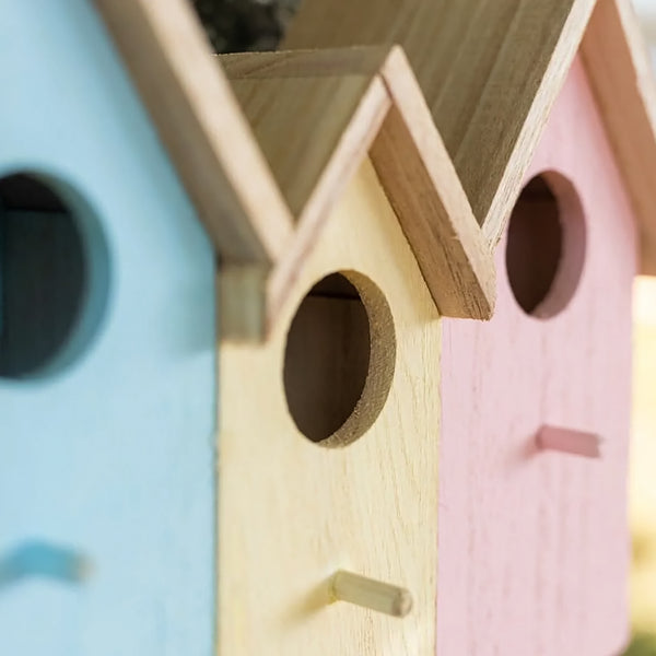 Three small house-shaped birdhouses in pastel blue, natural wood, and pink, shown close up with circular entry holes and small perches.