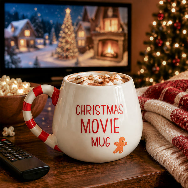 A white ceramic mug with a red-and-white striped handle and red text reading 'CHRISTMAS MOVIE MUG' sits on a wooden table in a cozy holiday setting. A TV in the background shows a snowy village scene, with a Christmas tree, popcorn bowl, remote control, and knitted blanket nearby.