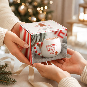A person holds a gift box containing a white ceramic Christmas movie mug with red text and a candy cane handle, set against a cozy holiday background with blurred lights and pine branches.