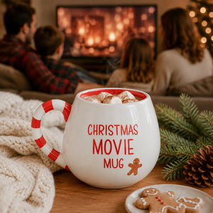 A white ceramic mug with a red and white candy cane handle sits on a wooden table in a cozy living room setting. The mug is filled with marshmallows and hot chocolate, and the words 'CHRISTMAS MOVIE MUG' are printed on the front in red, alongside a small gingerbread man graphic.