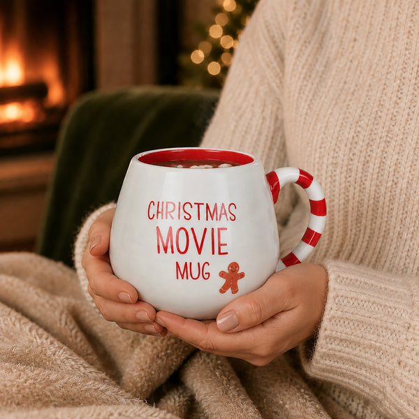 A person in a cream knit sweater holds a white ceramic mug with red text reading 'CHRISTMAS MOVIE MUG' and a red gingerbread figure graphic. The mug has a red handle and red interior, and the background shows a cozy indoor scene with a fireplace and warm lights.