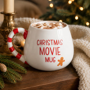 A white ceramic Christmas movie mug with a red and white candy cane handle, filled with a hot cocoa drink topped with marshmallows. The mug features the text 'CHRISTMAS MOVIE MUG' in red and a small gingerbread man graphic.